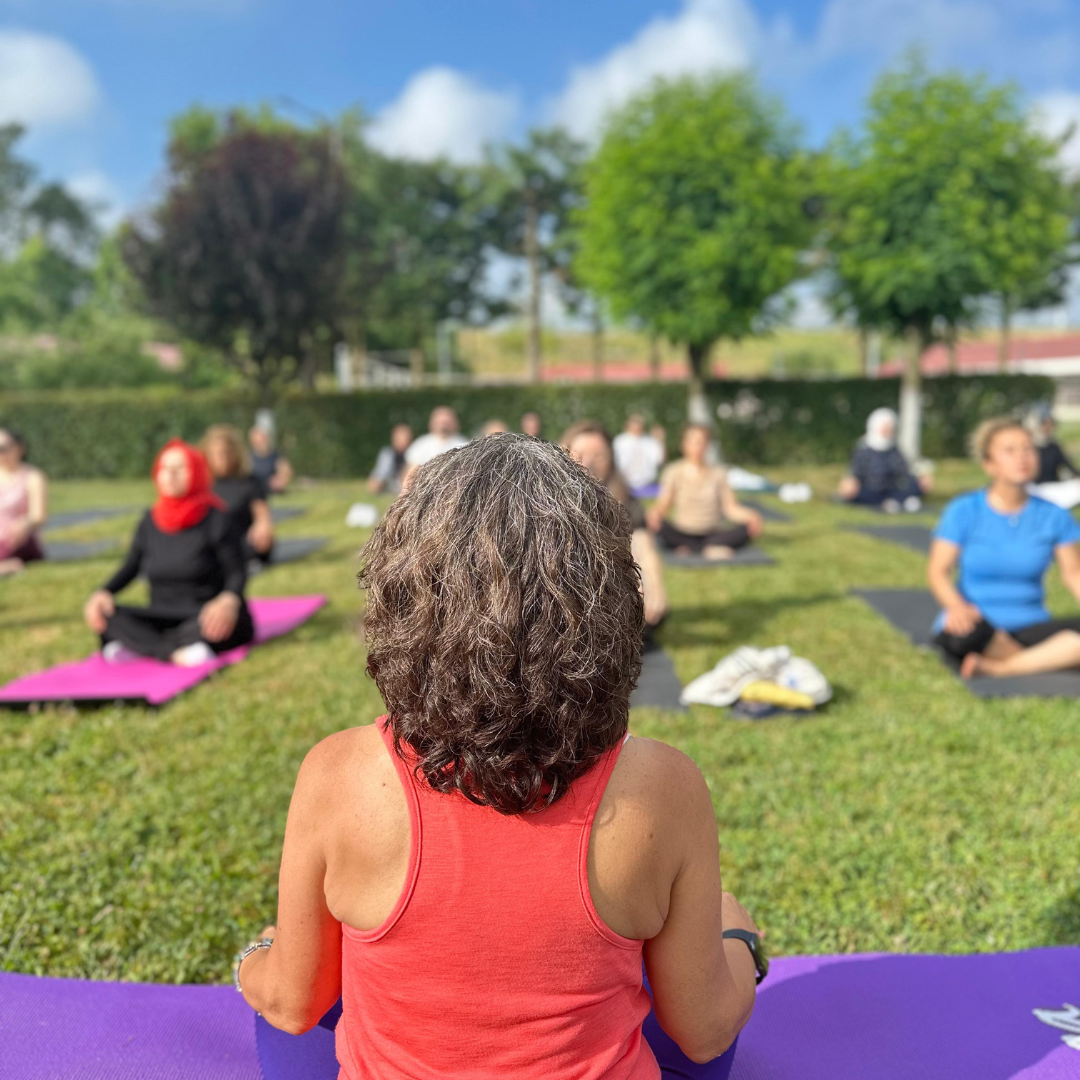 Figen Tekkol sırtı kameraya dönük, çimenler üzerinde bir grup insana meditasyon yaptırıyor.