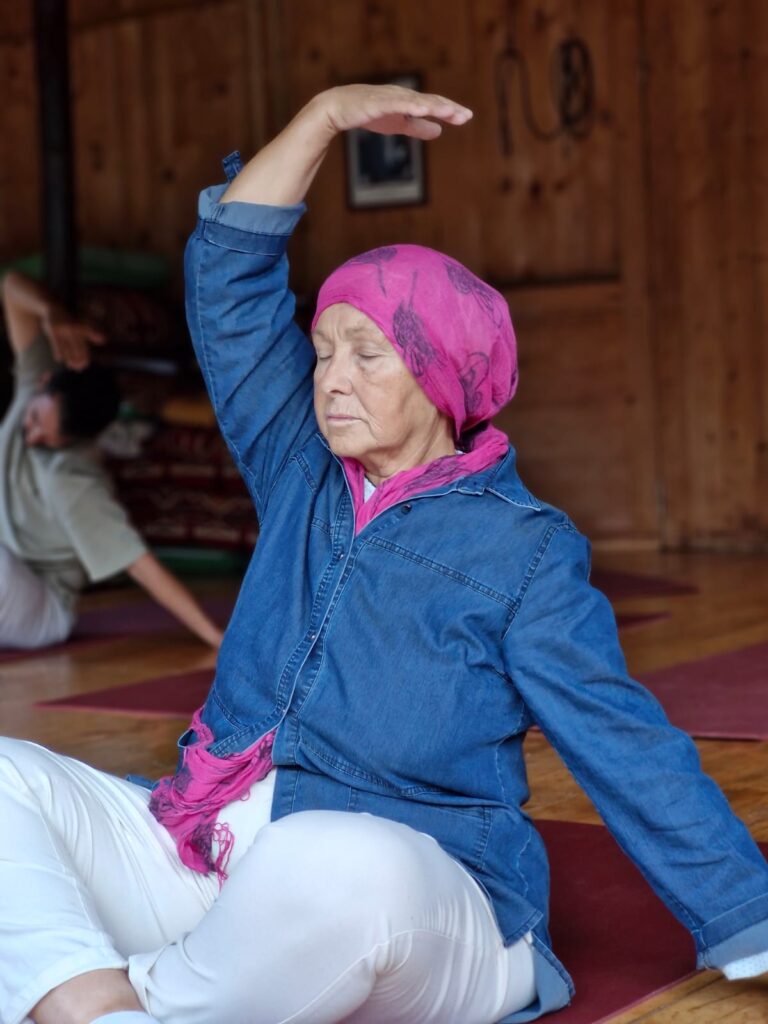 Figen Tekkol'un 50 yaş üstü öğrencisi, ahşap bir yoga stüdyosunda, pembe bandana takarak bir yoga pozu sırasında nazik bir şekilde beden farkındalığına odaklanırken.