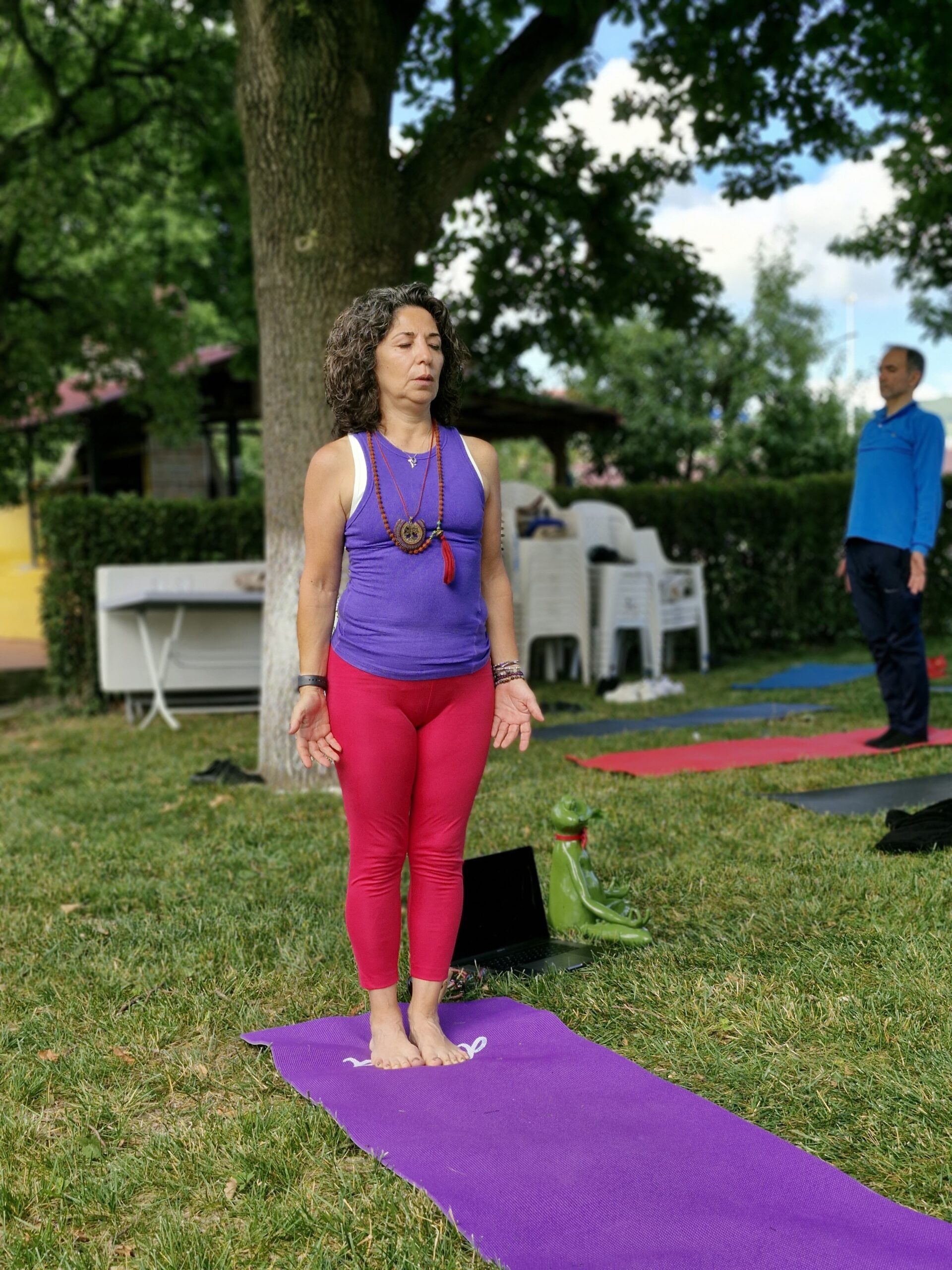 Figen Tekkol, açık alanda huzurlu bir ortamda yoga matı üzerinde ayakta gözleri kapalı tadasana yaparken. Yanında meditasyon yapan kaplumbağa heykeli ve laptop bulunuyor, sol tarafında erkek bir öğrencisi var.